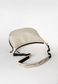 Beige Umhängetasche aus glattem, gepolstertem Stoff, mit verstellbarem schwarzen Tragegurt und einem Reißverschluss entlang der oberen Kante.