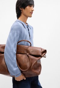 Sac de sport en cuir marron avec un rabat supérieur et des poignées doubles ; présente une texture lisse et un grain subtil. Le modèle porte une chemise à motifs bleu clair.