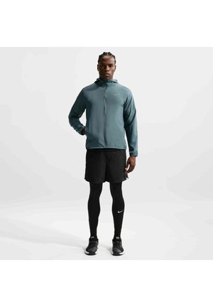 Mand stående opret iført en blågrøn lynlåshættetrøje, sorte shorts, sorte leggings og sorte løbesko på hvid baggrund.