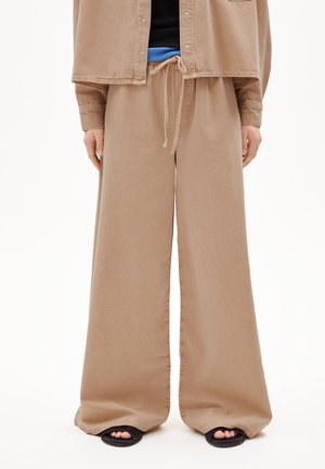 Weite Hosen aus einem hellbraunen Stoff mit einem Kordelzugbund, glatter Textur und seitlichen Schlitzen, kombiniert mit offenen schwarzen Sandalen.