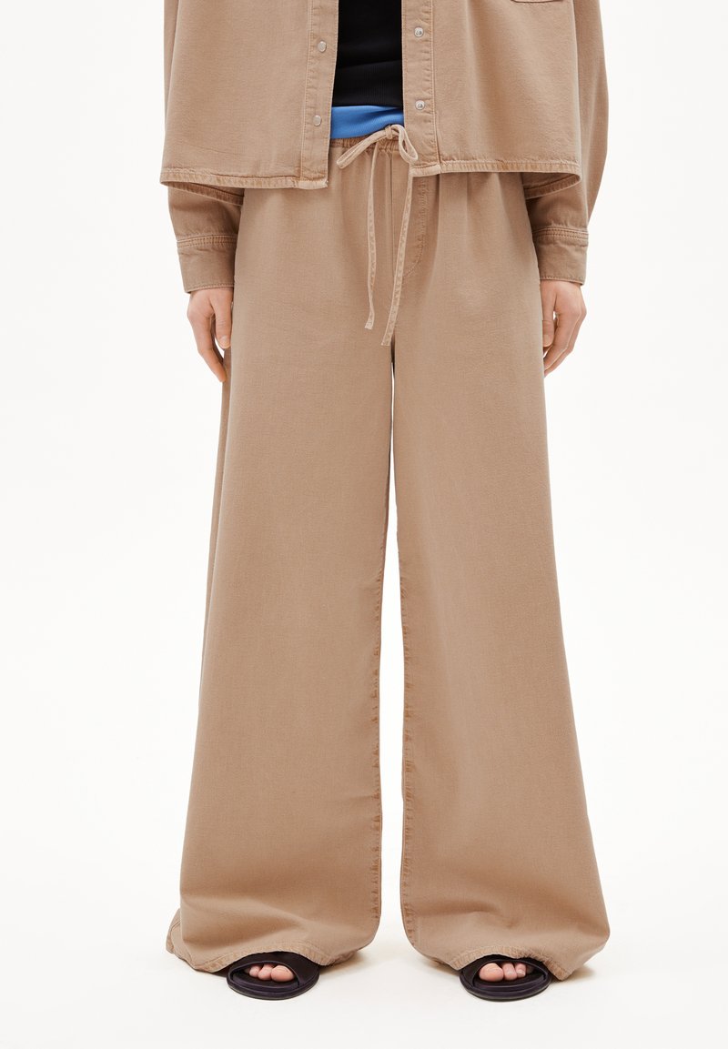 Pantalons larges en tissu brun clair, dotés d'une taille à cordon de serrage, d'une texture lisse et d'ouvertures latérales, associés à des sandales noires à bout ouvert.