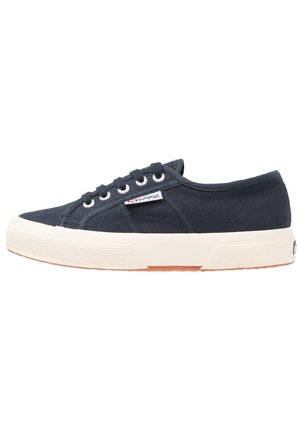 Zapatilla baja de lona negra con suela de goma blanca, cierre de cordones en la parte delantera y pequeña etiqueta de la marca Superga en el lateral.