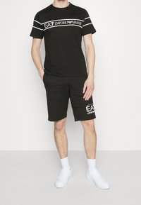Svart bomulls t-shirt med vita ränder och "EA7 Emporio Armani"-logotyp, i kombination med svarta shorts med en logotyp på sidan och vita sneakers.