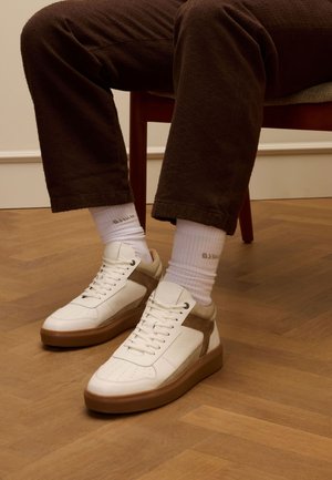 Witte leren sneakers met een tan suede accent, platte veters en een dikke bruine rubberen zool. Draagt met witte sokken en een bruine gestructureerde broek.