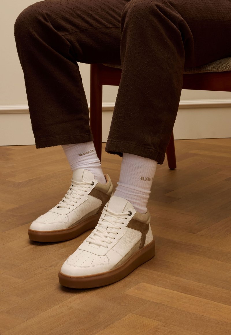 Witte leren sneakers met een tan suede accent, platte veters en een dikke bruine rubberen zool. Draagt met witte sokken en een bruine gestructureerde broek.