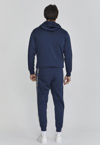 Marineblauwe hoodie met een trekkoord en lange mouwen; bijpassende broek heeft zijstrepen. Soepele textuur met elastische manchetten en tailleband.