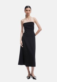 Robe noire sans bretelles avec un corsage ajusté et une jupe évasée arrivant aux genoux. Tissu lisse avec une texture douce. Mannequin portant des escarpins noirs.