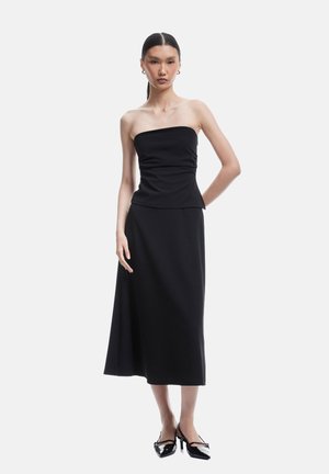 Robe noire sans bretelles avec un corsage ajusté et une jupe évasée arrivant aux genoux. Tissu lisse avec une texture douce. Mannequin portant des escarpins noirs.