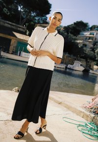 Femme debout près de l'eau portant un haut en maille à manches courtes blanc avec des bordures noires, une jupe noire et des sandales à talons noires.