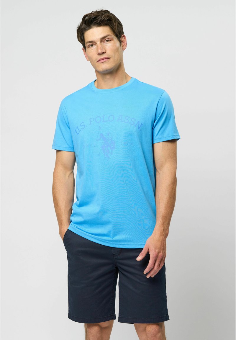 Lyseblå T-shirt i bomuld med korte ærmer, med et lilla U.S. Polo Assn. logo og design på fronten. Bæres med marineblå shorts.