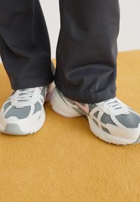 Zapatillas blancas y gris claro con paneles de malla y acentos en rosa; combinadas con pantalones negros, sobre un fondo amarillo.