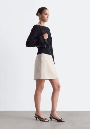 Femme portant un haut noir à manches longues, une jupe beige et des sandales brunes à petit talon, tenant un petit sac noir sur l'épaule.