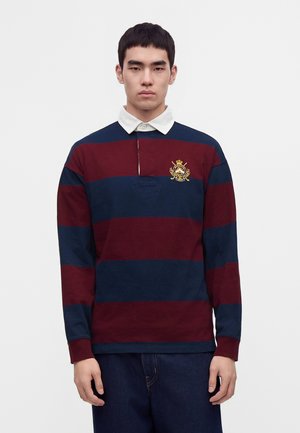 Homme portant un maillot de rugby rayé bleu marine et bordeaux avec un col blanc et un écusson brodé sur la poitrine gauche, debout devant un fond uni.