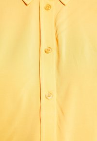 Chemise jaune à col avec une patte de boutonnage à l'avant comportant trois boutons jaunes visibles.