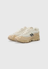 Scarpe sportive beige e crema realizzate con materiali in rete e suede, con punta arrotondata, suole testurizzate e un logo nero prominente sul lato.