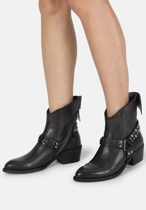 Bottes noires en cuir avec fermetures éclair latérales, brides cloutées et talons carrés bas, portées sur des jambes nues sur un fond blanc uni.