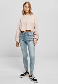 Donna che indossa un cropped sweatshirt di un rosa pallido, jeans skinny azzurri e sneakers bianco e nere, in piedi contro uno sfondo grigio uniforme.