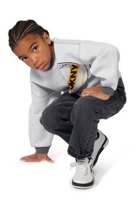 Sweatshirt gris avec logo noir et jaune, associé à un pantalon gris foncé et des baskets blanches avec des accents noirs. Le modèle est accroupi sur un genou.