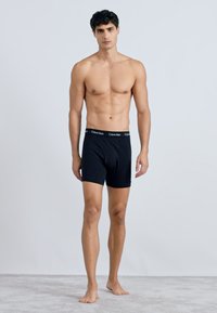 Boxer shorts de algodón negro con una cintura elástica que presenta un logo repetido. El modelo está descalzo sobre una alfombra clara.