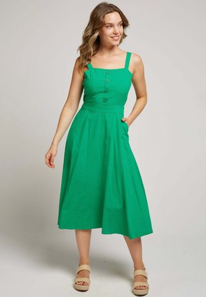 Femme souriante, debout avec une main dans la poche, portant une robe midi verte sans manches avec des boutons et des sandales compensées beiges.