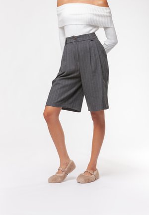 Pantaloni corti grigi a righe con vita alta, pieghe e chiusura a un bottone; abbinati a scarpe slip-on beige pelose.