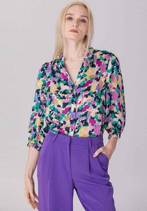 Femme aux longs cheveux blonds portant un chemisier coloré à motif abstrait avec des manches trois-quarts et un pantalon violet taille haute, main dans la poche.