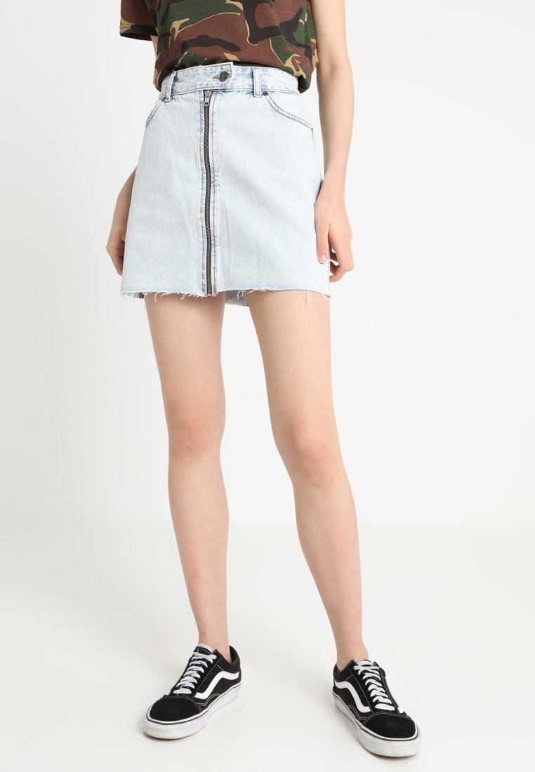 Personne portant un t-shirt camouflage, une mini-jupe en jean bleu clair avec fermeture éclair devant, et des baskets basses noires avec des lacets et semelles blanches.