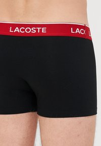 Boxer-briefs en coton extensible noir avec une ceinture rouge arborant le logo "LACOSTE" en blanc. Texture lisse et design ajusté avec des coutures minimales.