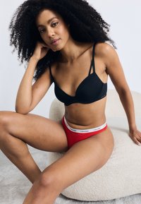 Svart polstret bh med tynne stropper og rød høyt midje undertøy med en hvit Calvin Klein-linning, satt mot en nøytral bakgrunn.
