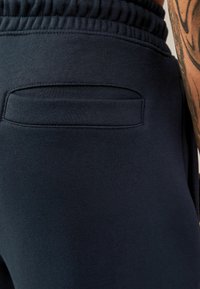 Joggers de un azul marino oscuro con un tejido suave, cinturilla elástica y un pequeño bolsillo trasero con un contorno cosido. Textura suave.
