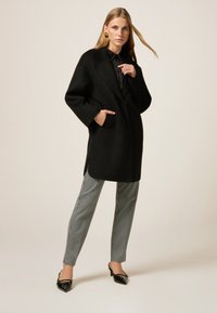 Cappotto in misto lana nero con maniche oversize, due tasche e spacchi laterali, abbinato a pantaloni a quadri houndstooth e décolleté a punta.