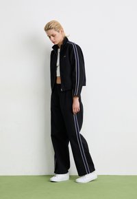 Svart zip-up jakke med hvite og marineblå stripe detaljer, kombinert med løse svarte bukser. Modellen har på seg en hvit crop top og hvite sneakers.