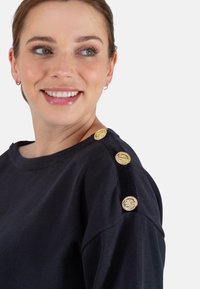 Schwarzes Sweatshirt mit Rundhalsausschnitt und Schulterakzenten, die mit goldenen Knopfdetails verziert sind. Der Stoff hat eine strukturierte Oberfläche.