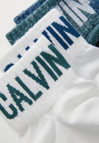 Drei Paar Socken flach ausgelegt: weiß mit dunkelgrüner Schrift, geprägter petrolfarbener MusterSocke und blaue Schrift auf einem strukturierten Hintergrund, Baumwollmischung.