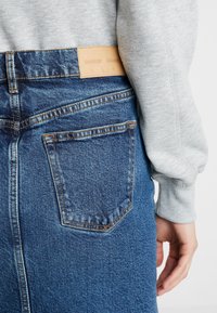 Jupe en denim bleu à taille haute, avec des détails de couture visibles et une étiquette de marque en cuir marron clair à l'arrière. Manches de sweat-shirt gris partiellement visibles.