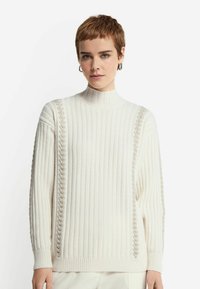 Creme ribbet striktrøje med høj krave og kabelstrikdetaljer langs siderne. Har lange ærmer og en afslappet pasform.