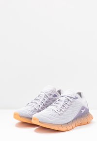 Para białych i lawendowych siateczkowych butów do biegania z pomarańczowymi podeszwami, logo Reebok po bokach i zawiązanymi sznurowadłami, prezentowanych na białej powierzchni.