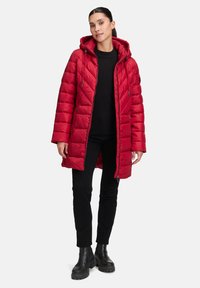 Rote Puffy-Jacke mit Kapuze, mit gestepptem Design, Frontreißverschluss und Kordeln. Trägt eine schwarze Bluse und Hose. Schwarze Stiefel.