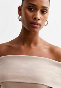 Ribbad off-shoulder topp i beige med en åtsittande design. Har en slät textur och en subtil lyster. Runda silverfärgade hoop örhängen.