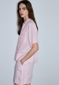 Mujer con cabello largo y castaño, vestida con una camiseta de manga corta rosa claro y pantalones cortos a juego, de pie con una mano en el bolsillo sobre un fondo gris.