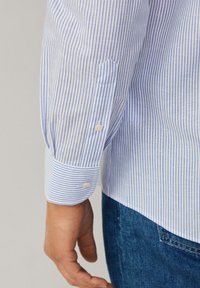 Blauw-wit gestreepte blouse met lange mouwen, voorzien van knoopmanchetten en een enkele zak op de linker mouw. Katoenen stof.