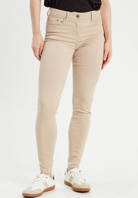 Jean skinny beige en tissu extensible, avec des poches avant et une fermeture à bouton, associés à des baskets blanches avec des accents marron.