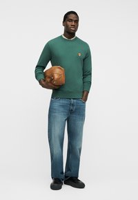 Homme en sweat-shirt vert avec logo de renard, jean bleu, baskets noires, tenant une pochette en cuir marron sur fond blanc.
