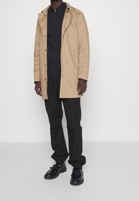 Beżowy wodoodporny płaszcz z kapturem, czarny sweter pod spodem, czarne spodnie cargo i czarne buty z frędzlami. Prosty, uproszczony design.