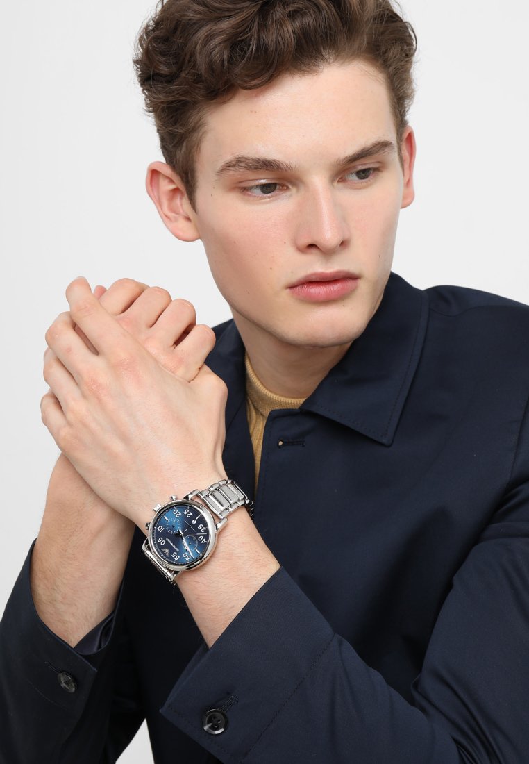 Reloj metálico plateado con una esfera azul, que presenta manecillas y marcadores de hora luminosos. Lleva puesto en una muñeca, complementado por una chaqueta oscura.