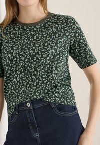 Femme portant une chemise à manches courtes à motifs verts et noirs rentrée dans un jean bleu foncé avec bouton et coutures visibles.