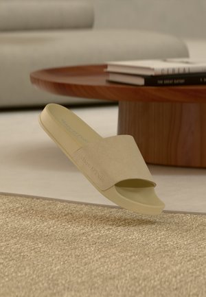 Beige liukusandaali leveällä hihnalla, jossa on kohokuvioitu "Marc O'Polo" -logo, kellumassa kuvioidun maton yläpuolella olohuoneessa, jossa on puinen sohvapöytä ja kirjoja.