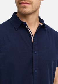 Camisa de botones de manga corta en color marino con un tejido texturizado, que presenta un cuello interior en contraste y botones en tonos similares.