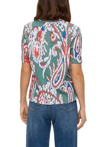 Krótki rękaw bluzki z zielonego materiału o plisowanej fakturze, z kolorowymi wzorami kwiatowymi w czerwonym, białym i niebieskim kolorze. Noszona z spodniami dżinsowymi o wysokim stanie.