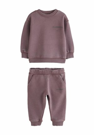 Young boy standing facing away, wearing mauve AllSaints sweatshirt and matching pants with white sneakers on gray background.